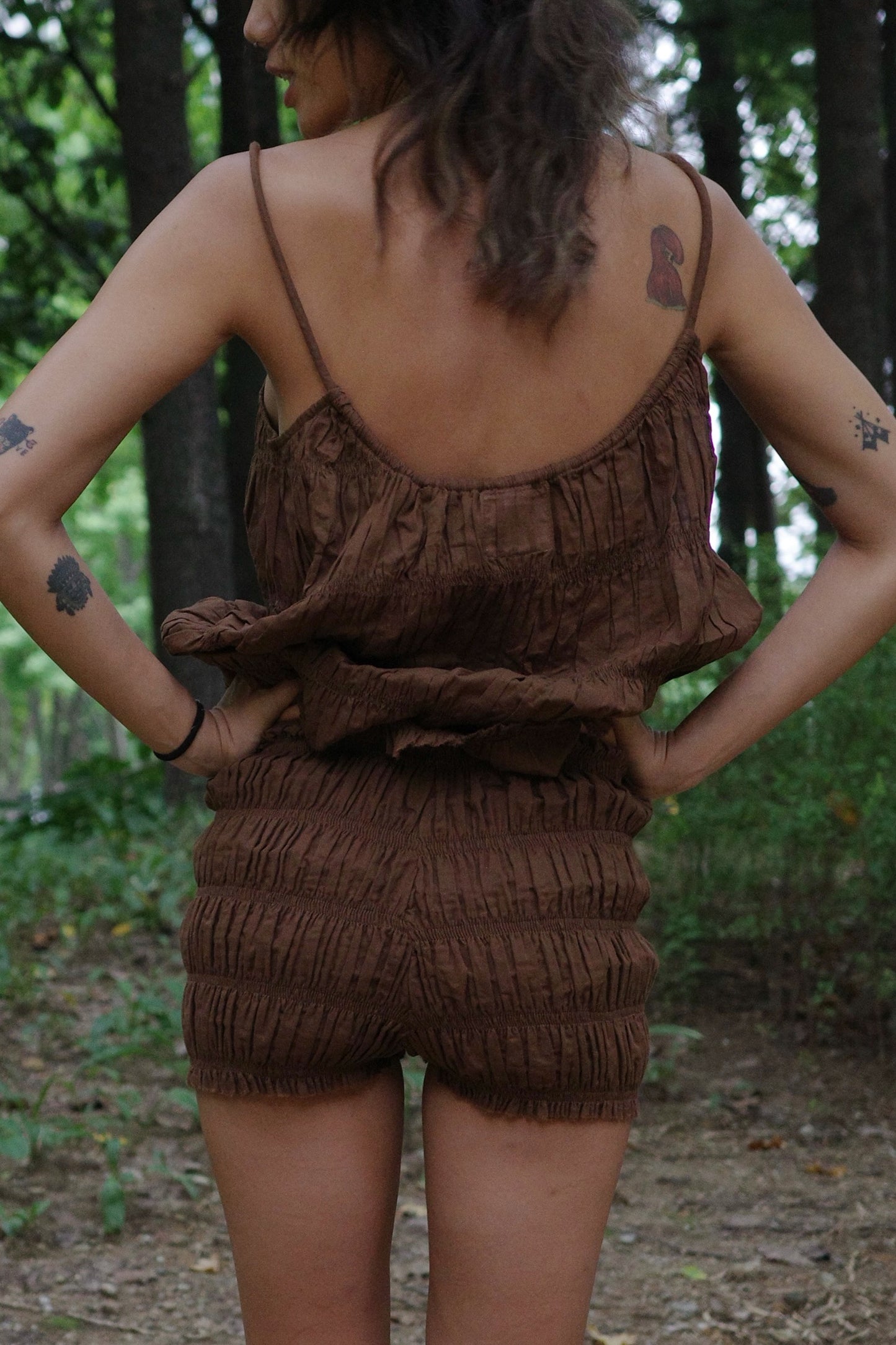 woman wearing a brown ruffled shorts and string top with tattoos on arms and back, standing in a forest