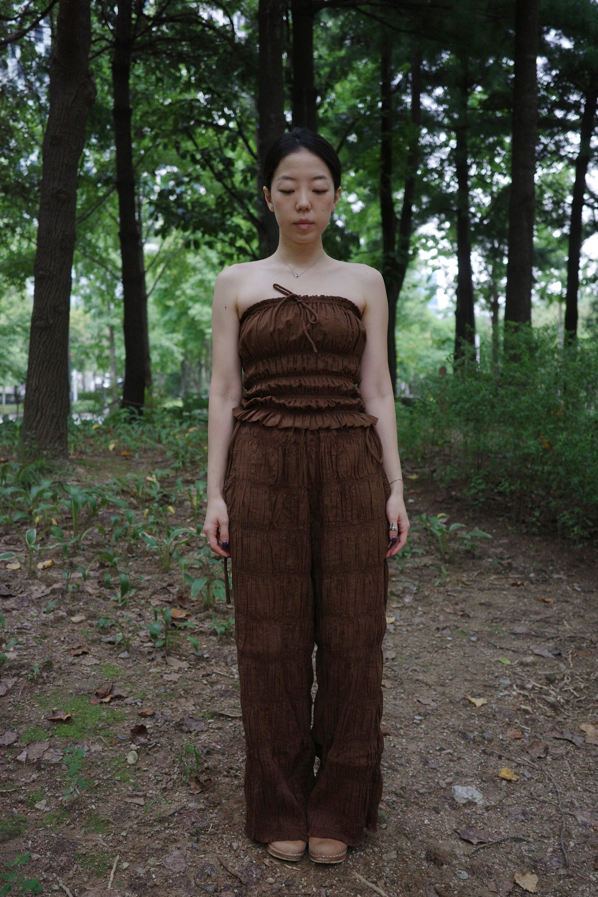 woman in a brown tube top and tie wide long pants standing in a forest