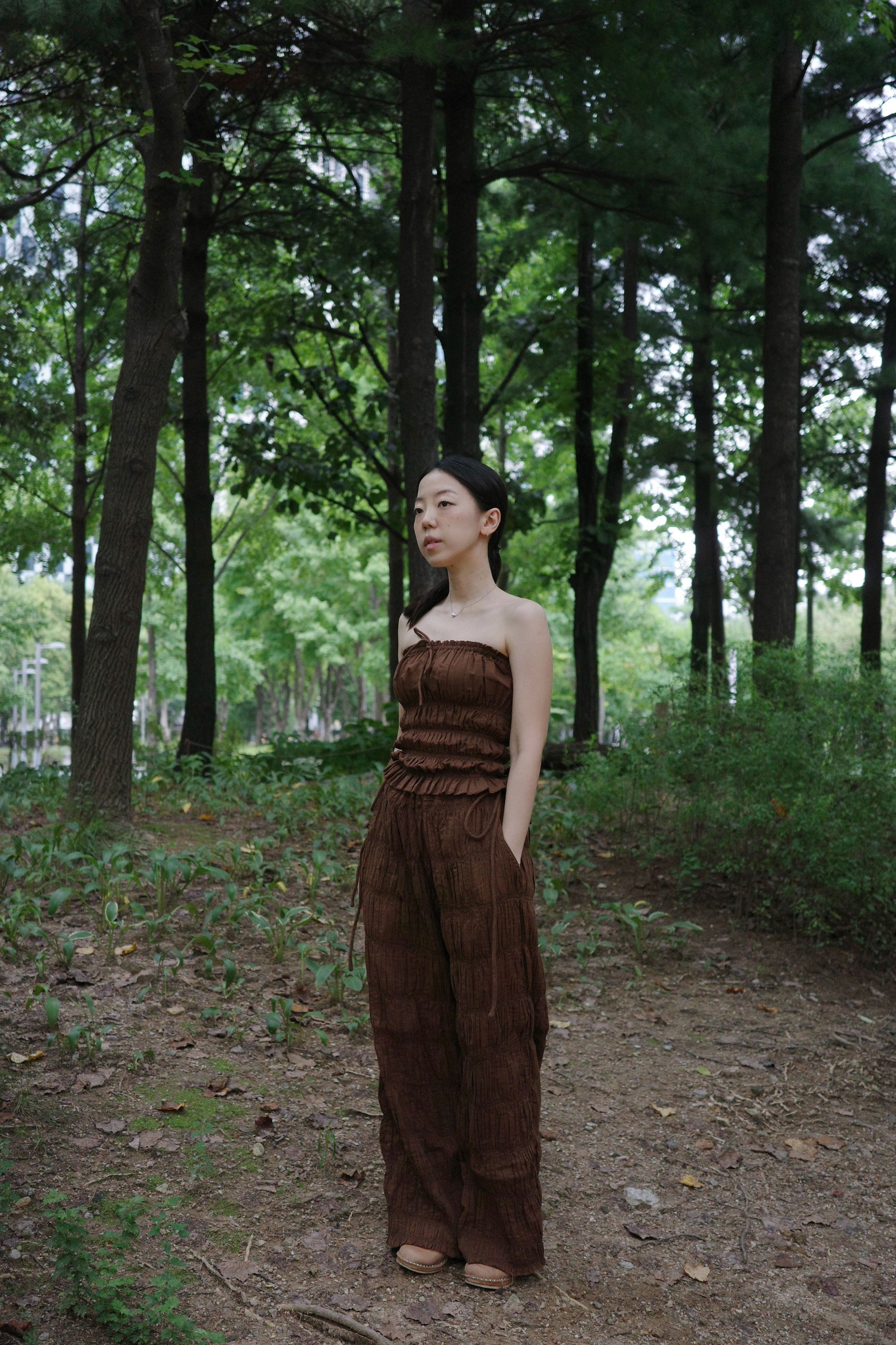 woman in a brown tube top and tie wide long pants standing in a forest