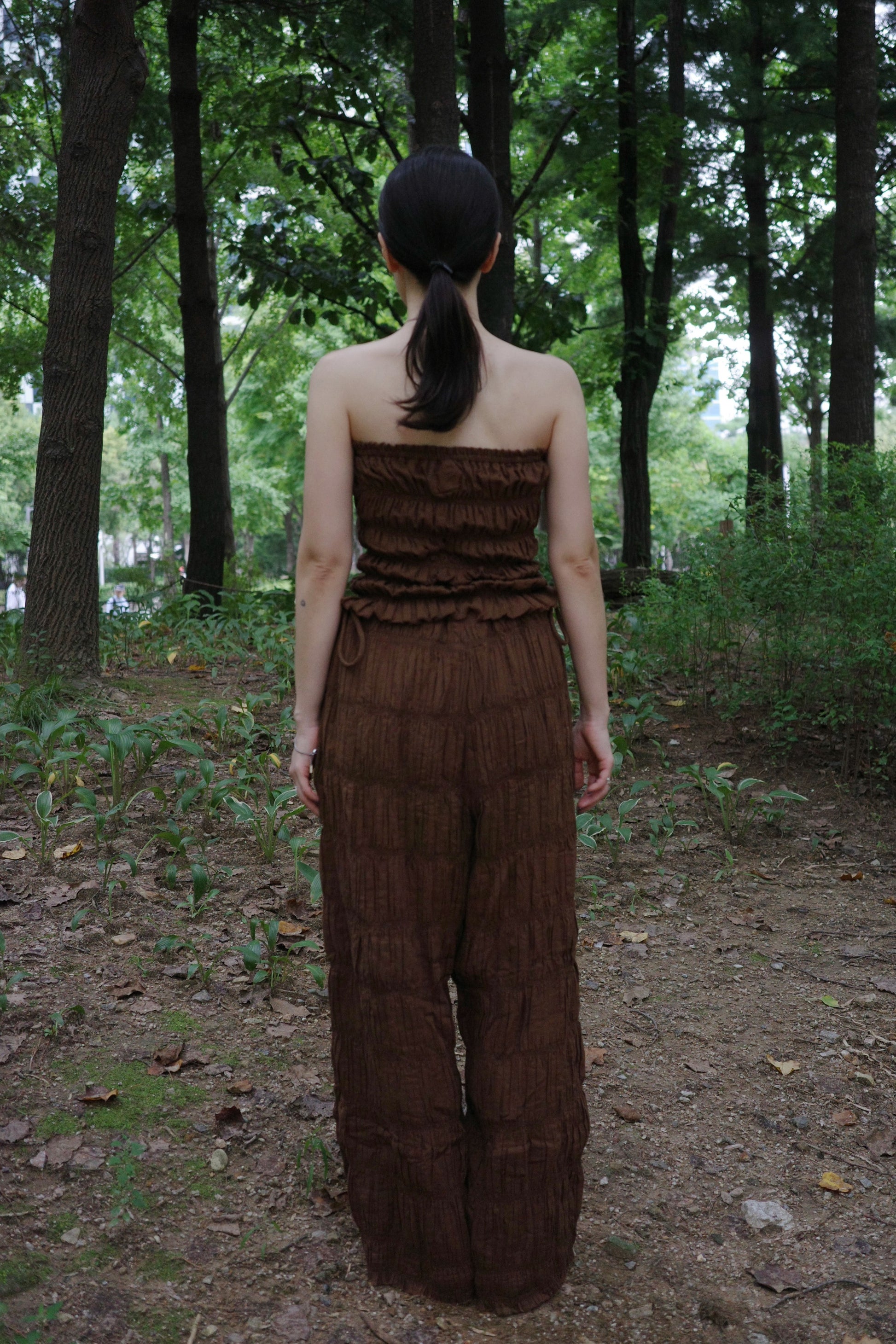 woman in a brown tube top and tie wide long pants standing in a forest