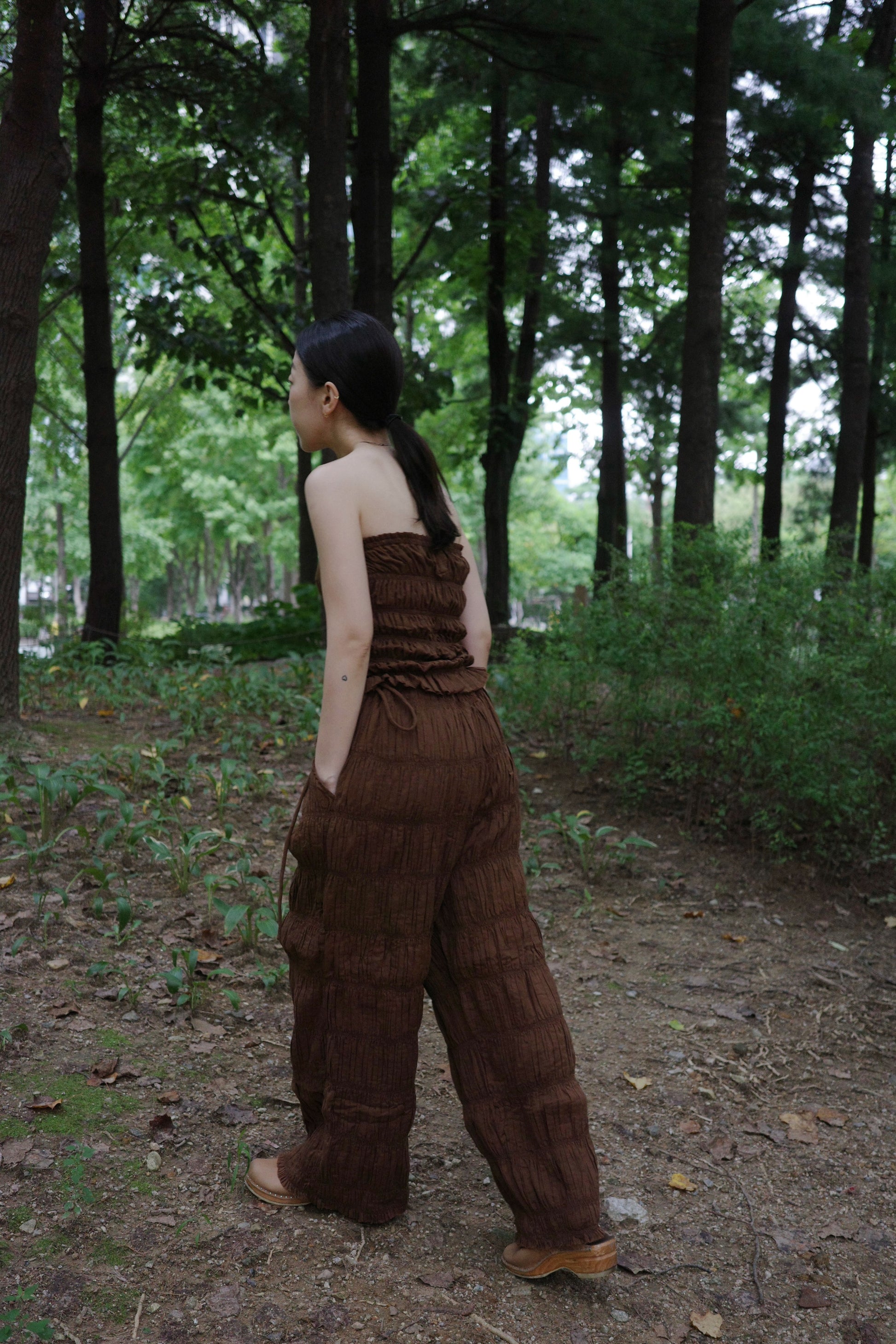 woman in a brown tube top and tie wide long pants walking through a forest