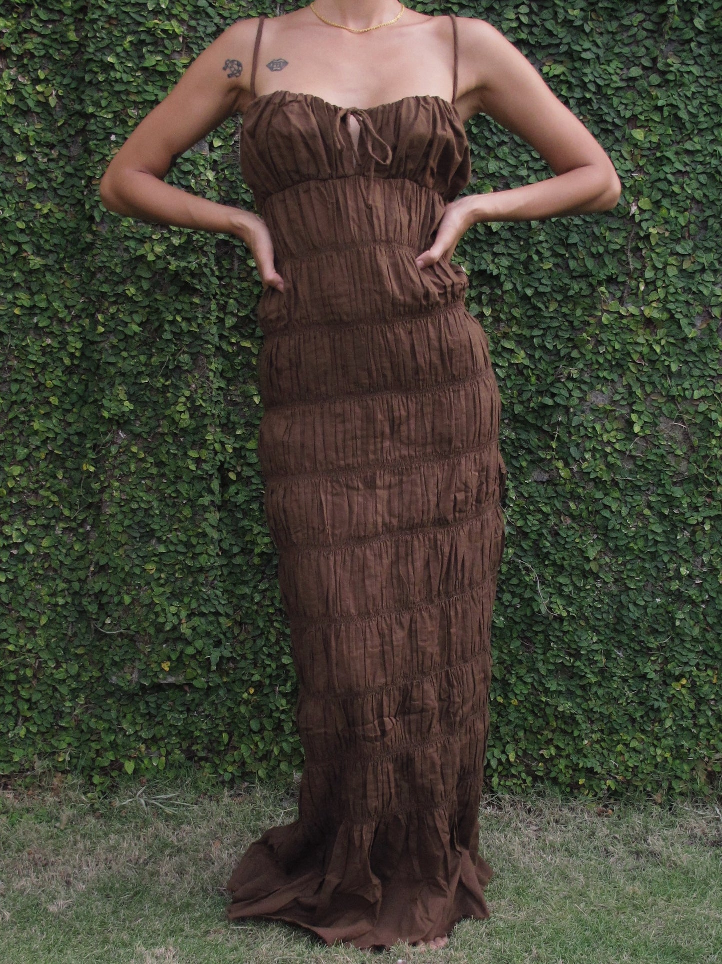 Woman wearing a brown ruffle dress against a green hedge backgroun