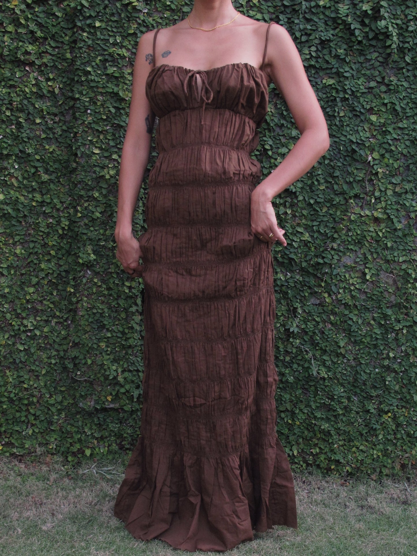 Woman wearing a brown ruffled dress against a green hedge background