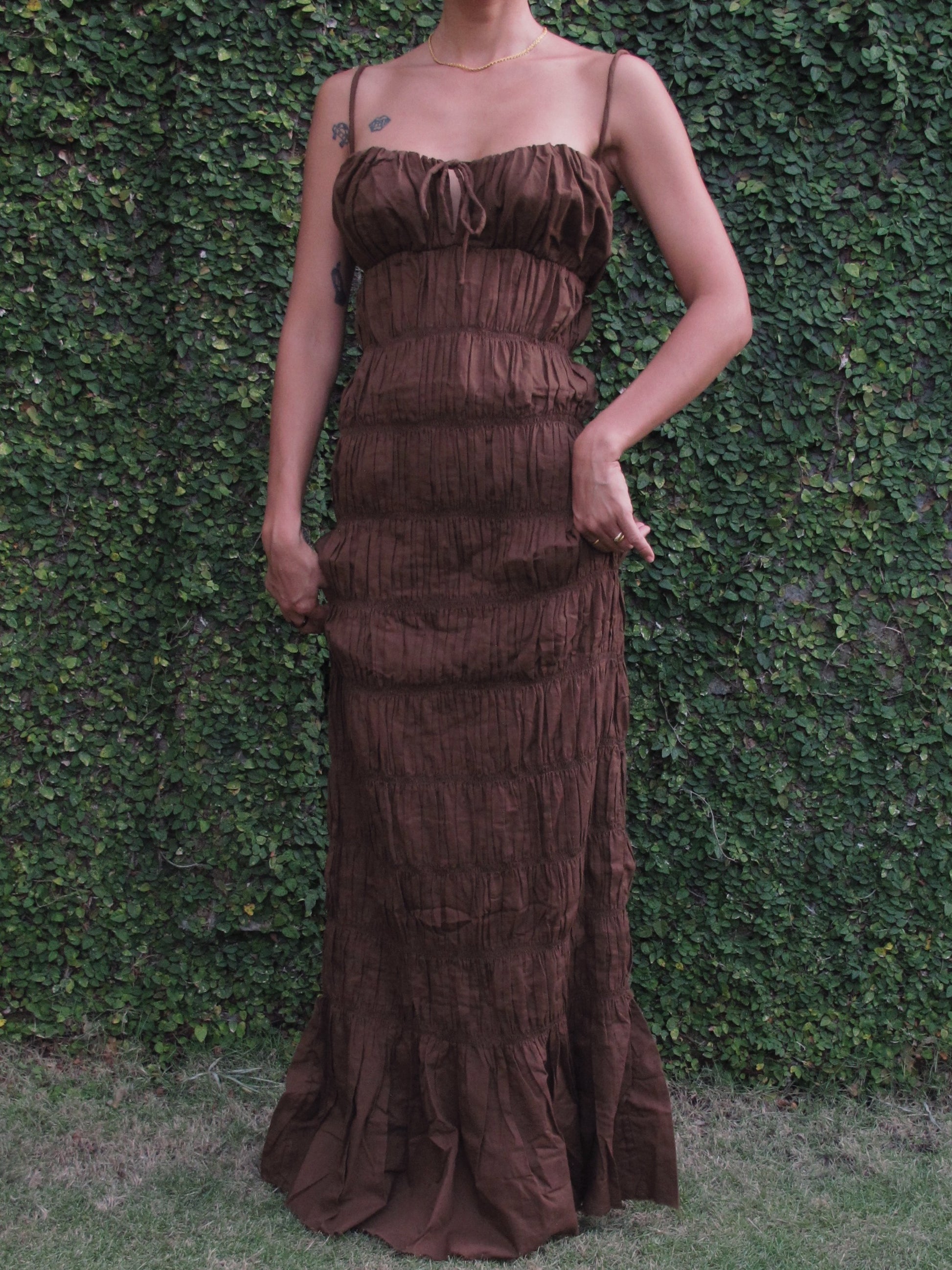 Woman wearing a brown ruffled dress against a green hedge background