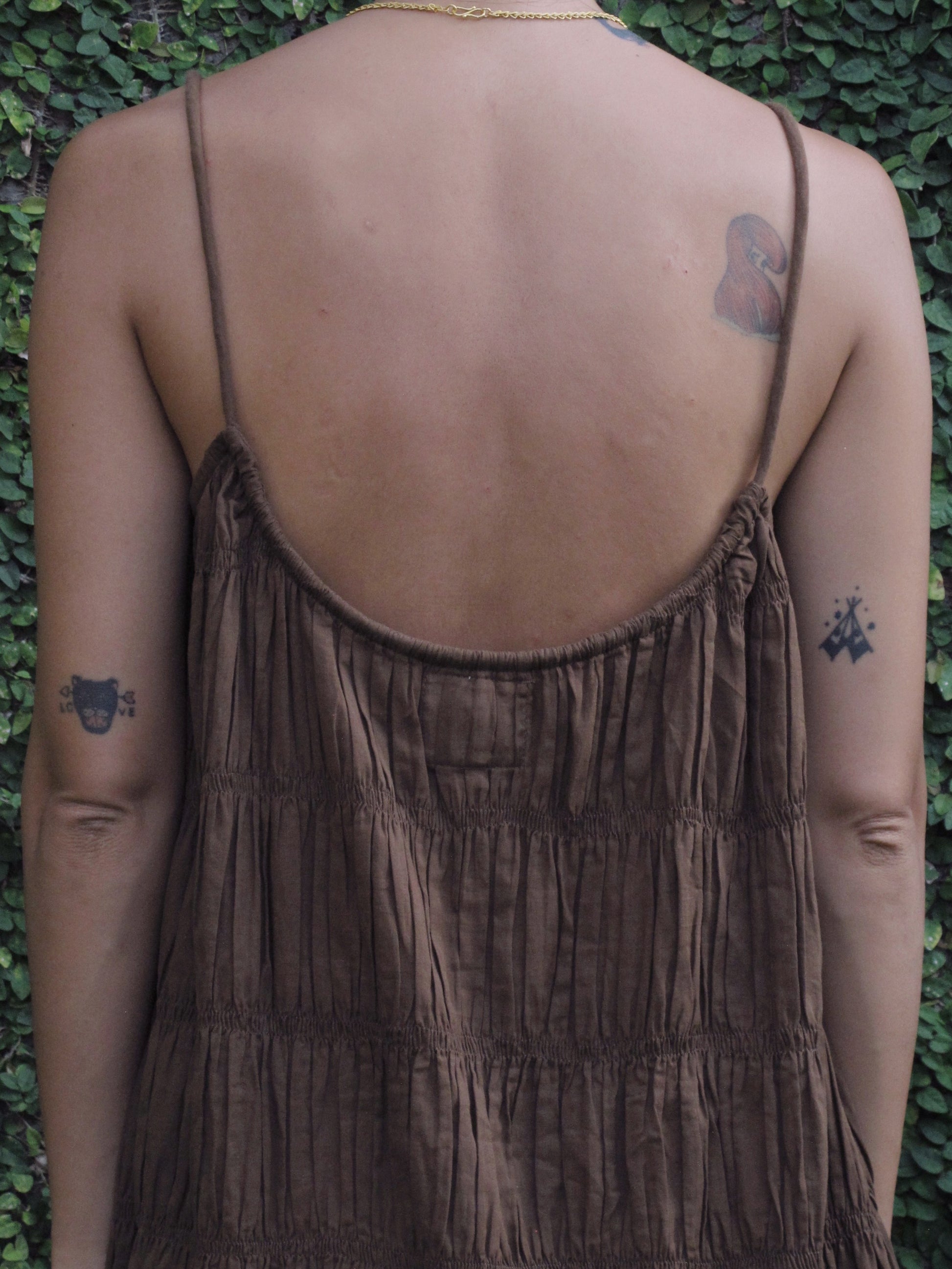 Brown sleeveless dress worn by a person with tattoos against a green leafy background