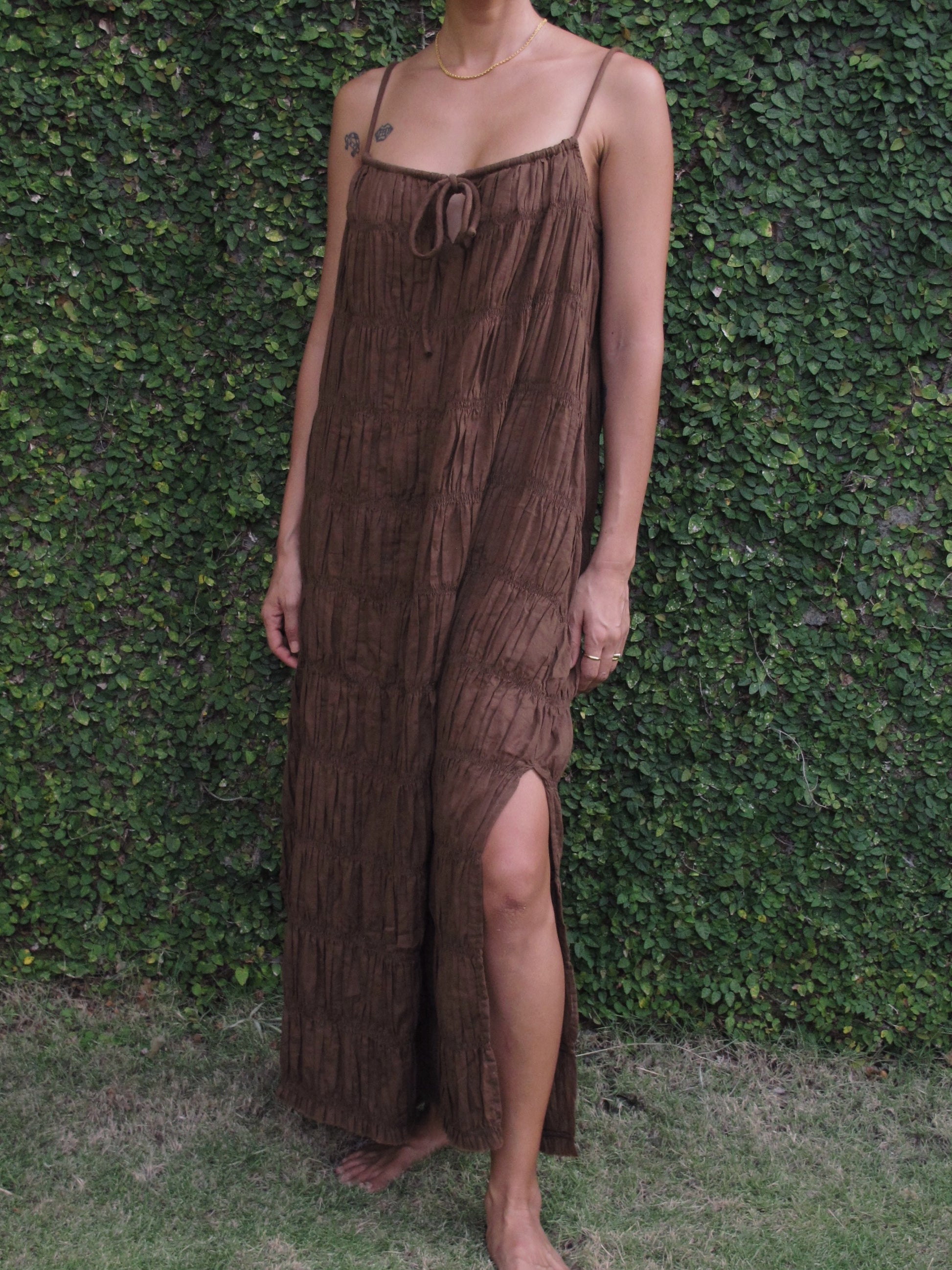 Person wearing a brown textured dress with a slit, standing against a green hedge.